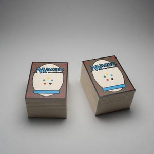 May include: Two stacks of Magic: The Gathering playing cards. The card backs are brown with a white oval design, the word "Magic" in blue, and colorful dots. The cards are stacked neatly on a light gray surface.