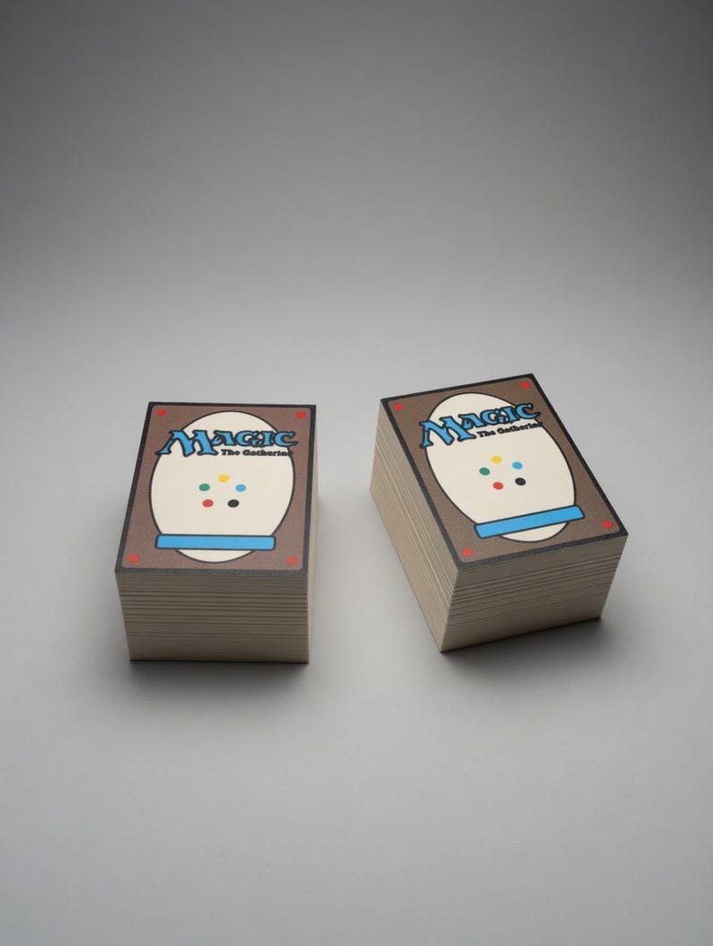 May include: Two stacks of Magic: The Gathering playing cards. The card backs are brown with a white oval design, the word "Magic" in blue, and colorful dots. The cards are stacked neatly on a light gray surface.