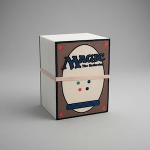 May include: A white, rectangular box of Magic: The Gathering trading cards, secured with a light brown rubber band. The box features a brown and cream-colored design with the word "Magic" in blue.