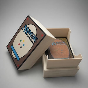 May include: A beige card storage box with the Magic: The Gathering logo on the lid. The box is open, revealing a stack of cards inside. The back of the cards are visible, with a brown border and colorful dots.