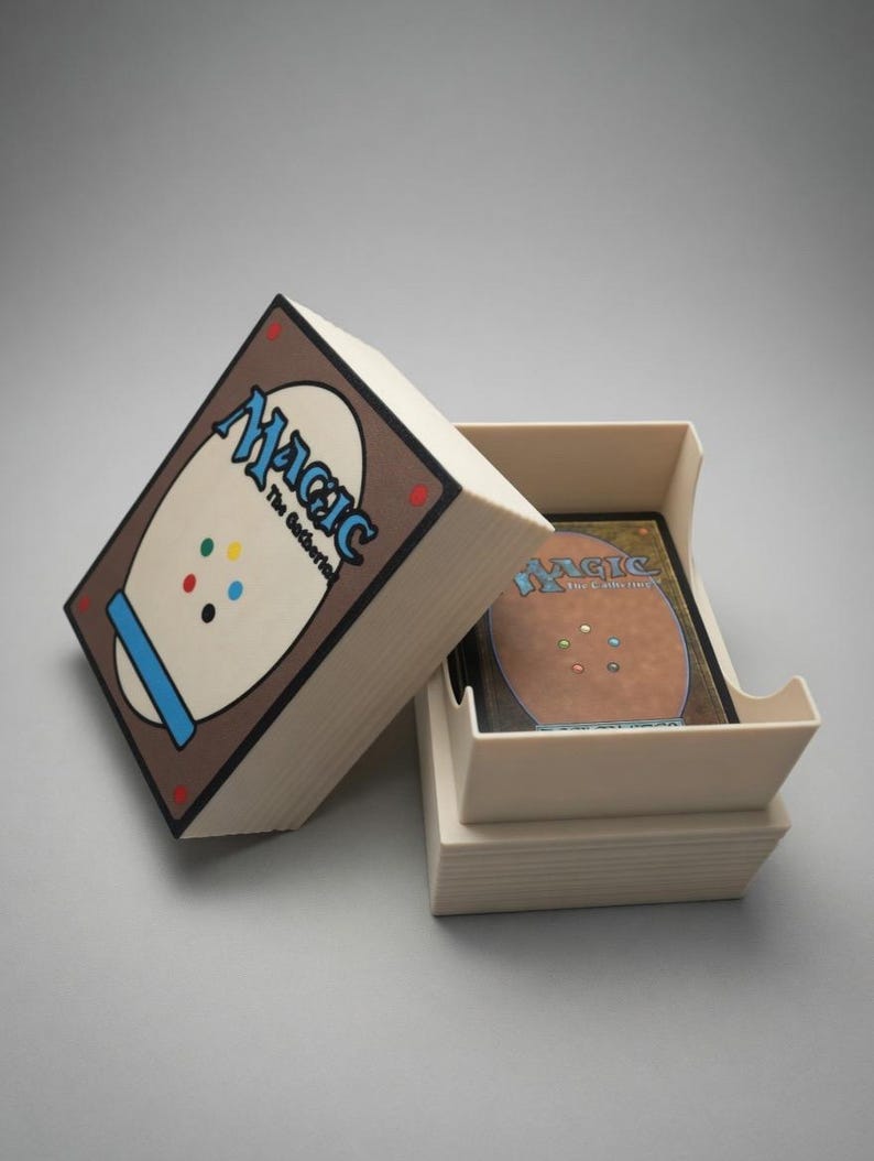 May include: A beige card storage box with the Magic: The Gathering logo on the lid. The box is open, revealing a stack of cards inside. The back of the cards are visible, with a brown border and colorful dots.