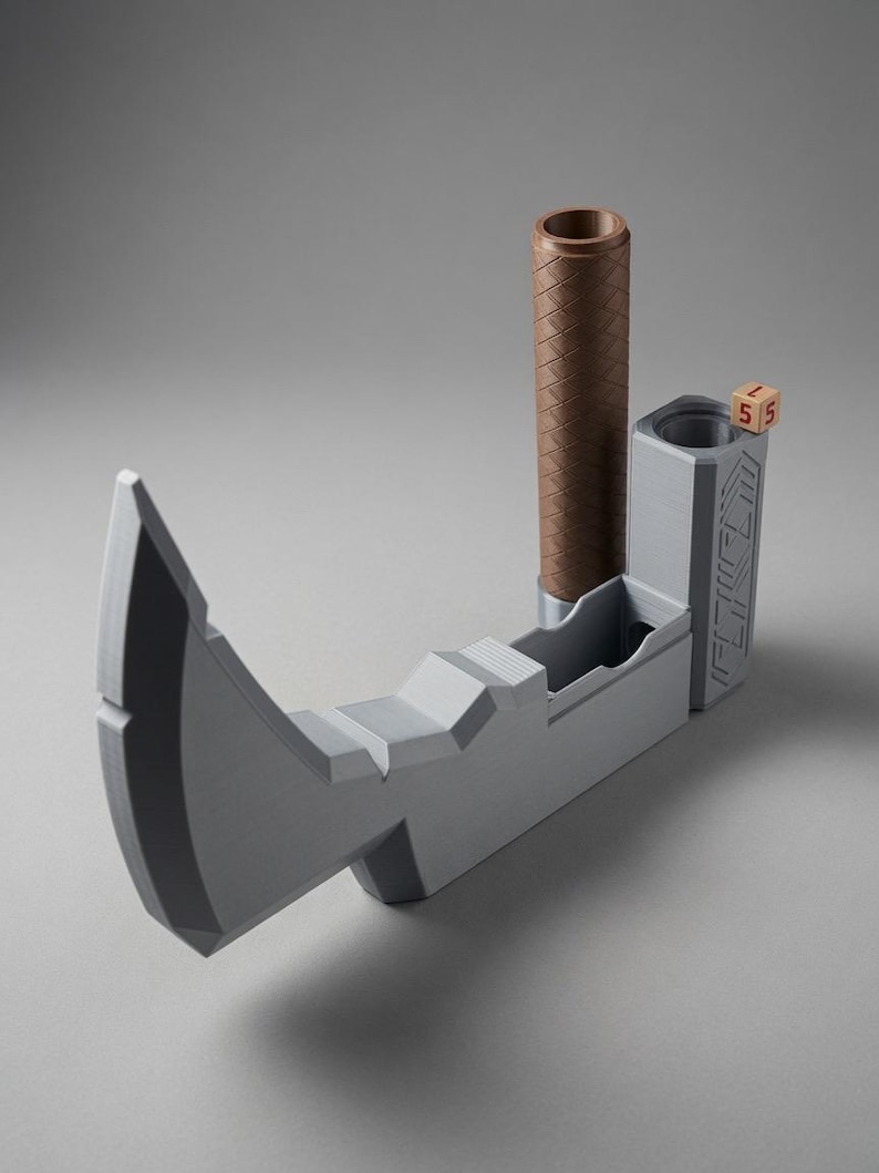 May include: A grey, axe-shaped dice tower featuring a brown, textured cylindrical handle and a vertical compartment for rolling dice. A small, light-coloured wooden die with the number 5 printed in red sits on the top edge of the tower structure.