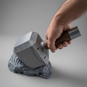 Op de afbeelding: Een zilverkleurige hamer met een rechthoekige kop voorzien van ingewikkelde, in reliëf aangebrachte knoopmotieven aan de zijkanten. Het handvat is bruin met een gestructureerd ruitpatroon en een zilveren knop. De hamer rust op een grijze rotsachtige basis.