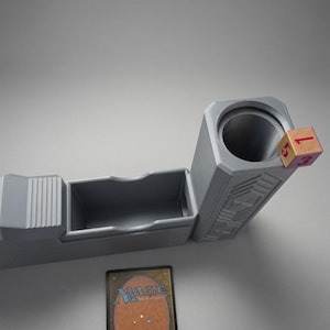 May include: A grey plastic deck box featuring a rectangular compartment for cards and a cylindrical tower for dice. A single wooden six-sided die with red numbers rests on the rim of the tower. A Magic: The Gathering trading card is placed below.