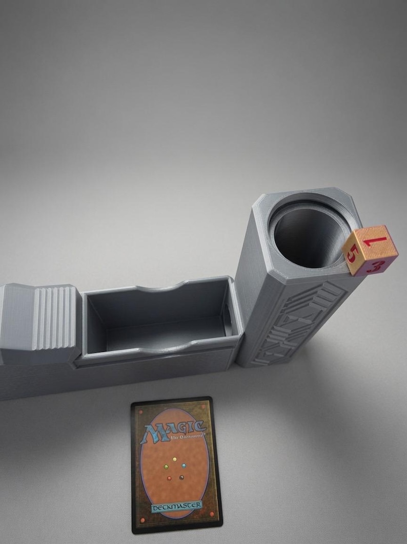 May include: A grey plastic deck box featuring a rectangular compartment for cards and a cylindrical tower for dice. A single wooden six-sided die with red numbers rests on the rim of the tower. A Magic: The Gathering trading card is placed below.