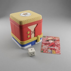May include: A square storage box designed with a red vest, blue shorts, and a yellow sash, topped with a textured straw hat lid featuring a skull emblem. Beside the box sits a white six-sided die and a rectangular trading card depicting a cartoon character.