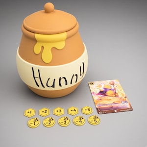May include: A tan honey pot with a lid, featuring a yellow drip design and the word Hunny printed on a cream band. Beside it are five yellow hexagonal tokens labeled +1 to +5 and five yellow circular tokens labeled -1 to -5 with a bee icon.