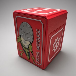 Puede incluir: Una caja de almacenamiento rectangular roja con un relieve de un personaje con máscara plateada y cabello rubio, con el texto ZECHS MERQUISE impreso verticalmente. El panel lateral muestra un emblema geométrico blanco sobre fondo rojo.