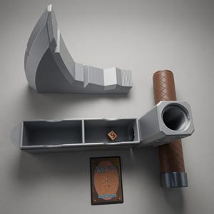 May include: A grey hammer-shaped deck box with a brown textured handle, featuring a detachable head that reveals an open storage compartment with two sections. Inside sits a single six-sided die showing the number five, next to a Magic The Gathering card.