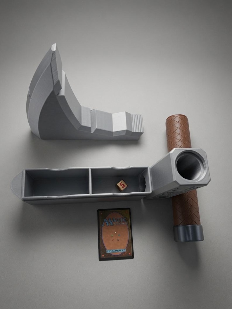May include: A grey hammer-shaped deck box with a brown textured handle, featuring a detachable head that reveals an open storage compartment with two sections. Inside sits a single six-sided die showing the number five, next to a Magic The Gathering card.