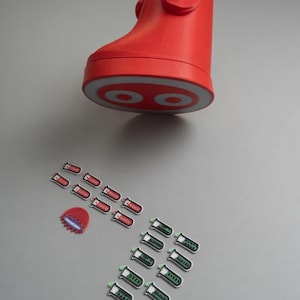 May include: A red, boot-shaped container with a white and red circular design on the sole, accompanied by a collection of small, rectangular stickers featuring red and green test tube graphics and a single red, jagged-edged circular sticker.
