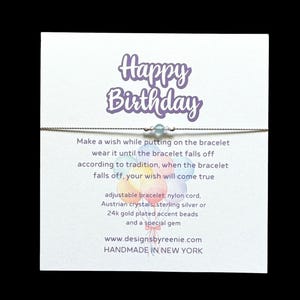 May include: A white card with the text "Happy Birthday" in purple lettering. The card has a silver string bracelet with a light blue gemstone bead. The card also includes instructions for making a wish and the text "www.designsbyreenie.com" and "HANDMADE IN NEW YORK".