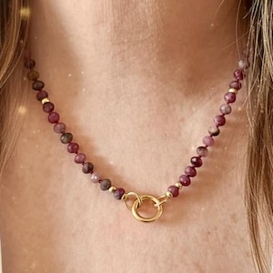 May include: A necklace featuring a strand of small, round, burgundy-colored beads. The necklace is accented with small gold-colored beads and a gold-colored double-ring clasp. The necklace is worn around a person's neck.