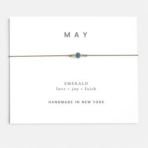 May include: A delicate bracelet on a white display card featuring the text MAY, EMERALD, love, joy, faith, and HANDMADE IN NEW YORK. The bracelet consists of a thin brown cord with a central green bead flanked by two small clear beads.