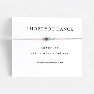 May include: A delicate bracelet with a light blue bead and a thin brown cord, presented on a white card. The card reads "I HOPE YOU DANCE" and "- BRACELET - wish . wear . believe". The text "HANDMADE IN NEW YORK" is also visible.