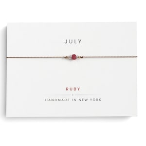 May include: A delicate brown cord bracelet featuring a small, faceted red ruby bead flanked by two tiny clear crystals, presented on a white display card printed with the text JULY, RUBY, and HANDMADE IN NEW YORK in black and red lettering.