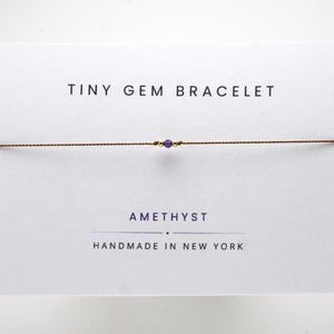 May include: A thin brown cord bracelet featuring a single small purple amethyst gemstone flanked by two tiny gold beads, displayed on a white card printed with the text TINY GEM BRACELET, AMETHYST, and HANDMADE IN NEW YORK.