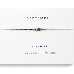 May include: A delicate bracelet featuring a single blue sapphire bead flanked by two small gold-tone beads and two clear crystals on a thin brown cord, presented on a white card labeled SEPTEMBER, SAPPHIRE, and HANDMADE IN NEW YORK.