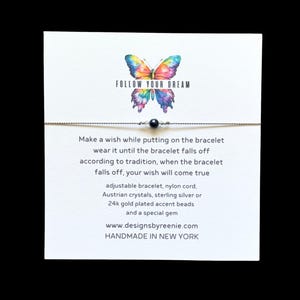 May include: A white card with a colorful butterfly illustration and the text "FOLLOW YOUR DREAM". The card also features a wish bracelet with a black bead and the text "Make a wish while putting on the bracelet wear it until the bracelet falls off according to tradition, when the bracelet falls off, your wish will come true adjustable bracelet, nylon cord, Austrian crystals, sterling silver or 24k gold plated accent beads and a special gem www.designsbyreenie.com HANDMADE IN NEW YORK"