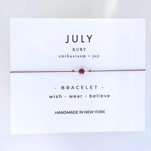 May include: A July ruby bracelet on a white card. The card reads "JULY RUBY enthusiasm • joy - BRACELET - wish • wear • believe HANDMADE IN NEW YORK". The bracelet has a red gemstone and gold accents.