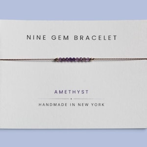 May include: A delicate bracelet featuring nine small, faceted purple amethyst beads and two tiny gold-toned beads on a thin brown cord. The bracelet is displayed on a white card printed with the text NINE GEM BRACELET, AMETHYST, and HANDMADE IN NEW YORK.