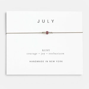 May include: A delicate bracelet featuring a single small red ruby bead centered on a thin brown cord. The bracelet is displayed on a white card printed with the text JULY, RUBY, COURAGE • JOY • ENTHUSIASM, and HANDMADE IN NEW YORK.