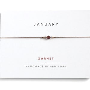 May include: A thin brown cord bracelet featuring a central dark red garnet bead flanked by two tiny clear crystals and two small gold-tone beads, displayed on a white card printed with the text JANUARY, GARNET, and HANDMADE IN NEW YORK.