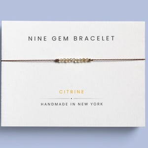 May include: A delicate bracelet featuring nine small, faceted yellow citrine gemstones strung on a thin brown cord. The bracelet is displayed on a white card with the text NINE GEM BRACELET, CITRINE, and HANDMADE IN NEW YORK printed in black and gold.