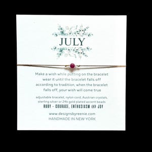 May include: A delicate July wish bracelet with a ruby bead, displayed on a white card. The card features floral accents and text that reads "JULY". The bracelet is made with a nylon cord and Austrian crystals. The card also includes the words "RUBY - COURAGE, ENTHUSIASM AND JOY".