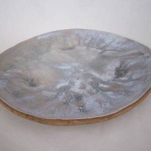 May include: An oval-shaped decorative tray with a light gray, marbled surface and a natural wood-toned edge. The tray's surface has a flowing, organic pattern with hints of white and brown.