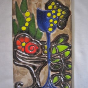 May include: A ceramic tile with a colorful floral design. The tile features a blue vase with yellow flowers, red flowers, and green leaves. The tile has a brown background.