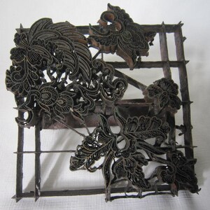 May include: A dark brown metal batik stamp featuring an intricate floral and leaf pattern. The tool consists of a square grid frame with raised metal strips shaped into detailed botanical motifs, designed for applying wax patterns to fabric.