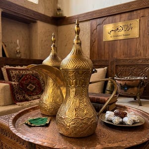 May include: Two gold-coloured coffee pots with intricate designs and long spouts are displayed on a copper tray. The tray also holds a small plate of wrapped dates and a green cloth. The background features a wooden wall with Arabic text and the words "Al Ibrahimi Bazaar".