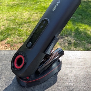 May include: Black electronic device with the word "ispire" printed on the side. The device has a red accent around the opening and base. Two black batteries are inserted into the base. The device is on a gray surface.