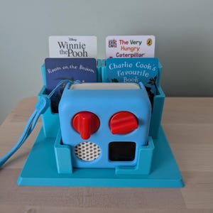 May include: A light blue Yoto audio player with two red control knobs, a white speaker grille, and a black display screen. It sits in a matching blue plastic stand holding four story cards: Winnie the Pooh, Room on the Broom, The Very Hungry Caterpillar, and Charlie Cook's Favourite Book.