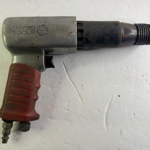 Può includere: Un martello pneumatico con corpo argentato, sezione anteriore nera e impugnatura rossa. Lo strumento presenta il logo "MATCO TOOLS" e una molla all'estremità. Progettato per uso industriale.