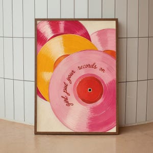 May include: A framed wall art print featuring a stack of colorful vinyl records in shades of pink, yellow, and red. The central pink record displays the handwritten text "girl put your records on" in a dark red script font against a light background.