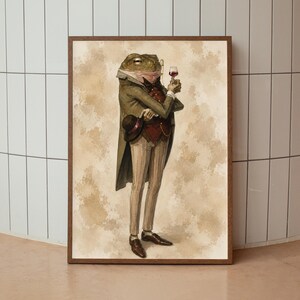 May include: A framed wall art print depicting an anthropomorphic frog dressed in a formal olive green tailcoat, a dark red vest, and light pinstriped trousers. The frog wears a monocle, holds a small glass of red wine, and carries a black top hat.