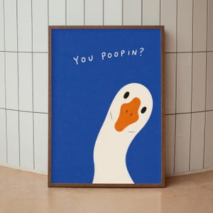 May include: A framed wall art print featuring a white goose with an orange beak on a solid blue background. The goose is positioned in the lower right corner, looking upward. The text "You Poopin?" is printed in white in the upper left area.