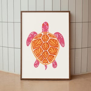 Puede incluir: Una lámina de arte mural enmarcada con una ilustración estilizada de una tortuga marina. La tortuga tiene la cabeza y las aletas de color rosa brillante con puntos blancos, y un caparazón naranja decorado con patrones de hojas simétricos.