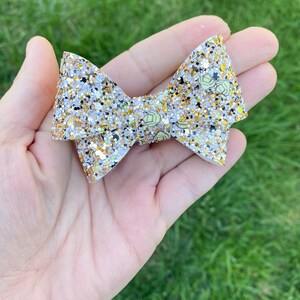 May include: A white and gold glitter bow with a volleyball pattern. The bow is tied in a knot and has a small loop at the top.