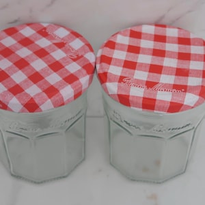 Könnte beinhalten: Zwei klare Gläser mit rot-weiß karierten Stoffdeckeln. Die Gläser haben eine sechseckige Form und sind leer. Auf den Deckeln steht "Rameau Masson". Die Gläser stehen auf einer weißen Oberfläche.