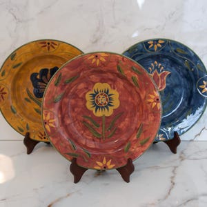 May include: Three decorative ceramic plates with floral designs. The plates are in shades of yellow, red, and blue, each featuring a central flower motif and a decorative border. They are displayed on small wooden stands.