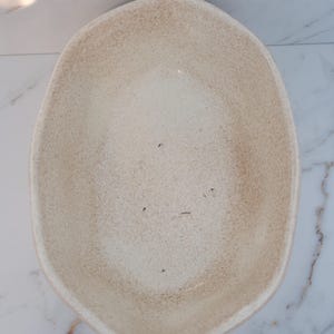 Pode incluir: Uma tigela de cerâmica bege, em forma oval, com uma textura salpicada. A tigela tem uma borda ligeiramente irregular e um interior liso. A cor neutra e o design simples tornam-na versátil.