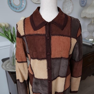 May include: A patchwork suede jacket in shades of brown, tan, and beige. The jacket features a collared neckline, button-down front, and crochet trim. The jacket is displayed on a mannequin.