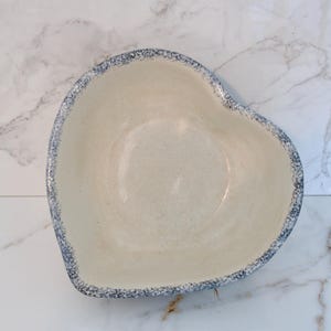 May include: Cream-colored, heart-shaped ceramic bowl with a speckled blue rim. The bowl has a smooth interior and a slightly textured exterior. Ideal for serving or as a decorative piece.