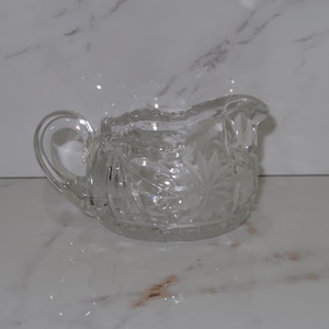 May include: Clear glass creamer with a handle and a starburst pattern. The creamer has a curved handle and a spout for easy pouring. The glass is clear and reflects light, creating a sparkling effect. The creamer is likely used for serving cream or milk.