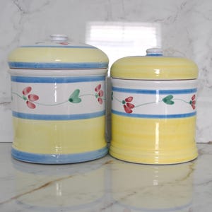 May include: Two ceramic canisters with lids. The canisters are yellow with blue and white stripes, and floral designs. The lids are yellow with a small white knob. The canisters are of different sizes.