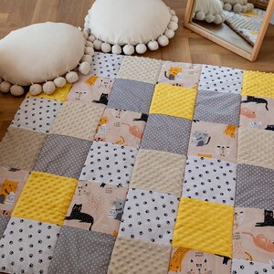 Puede incluir: Un edredón de patchwork con ilustraciones de gatos en beige, amarillo, gris y blanco. El edredón está sobre un suelo de madera con dos cojines redondos color crema con ribete de pompón. Un espejo está en el fondo.