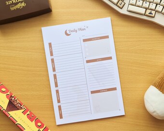 May include: A white daily planner with a brown and beige design, featuring the text "Daily Plan". The planner has sections for priorities, notes, and reminders. A chocolate box, a keyboard, and a plush coffee cup are also visible.